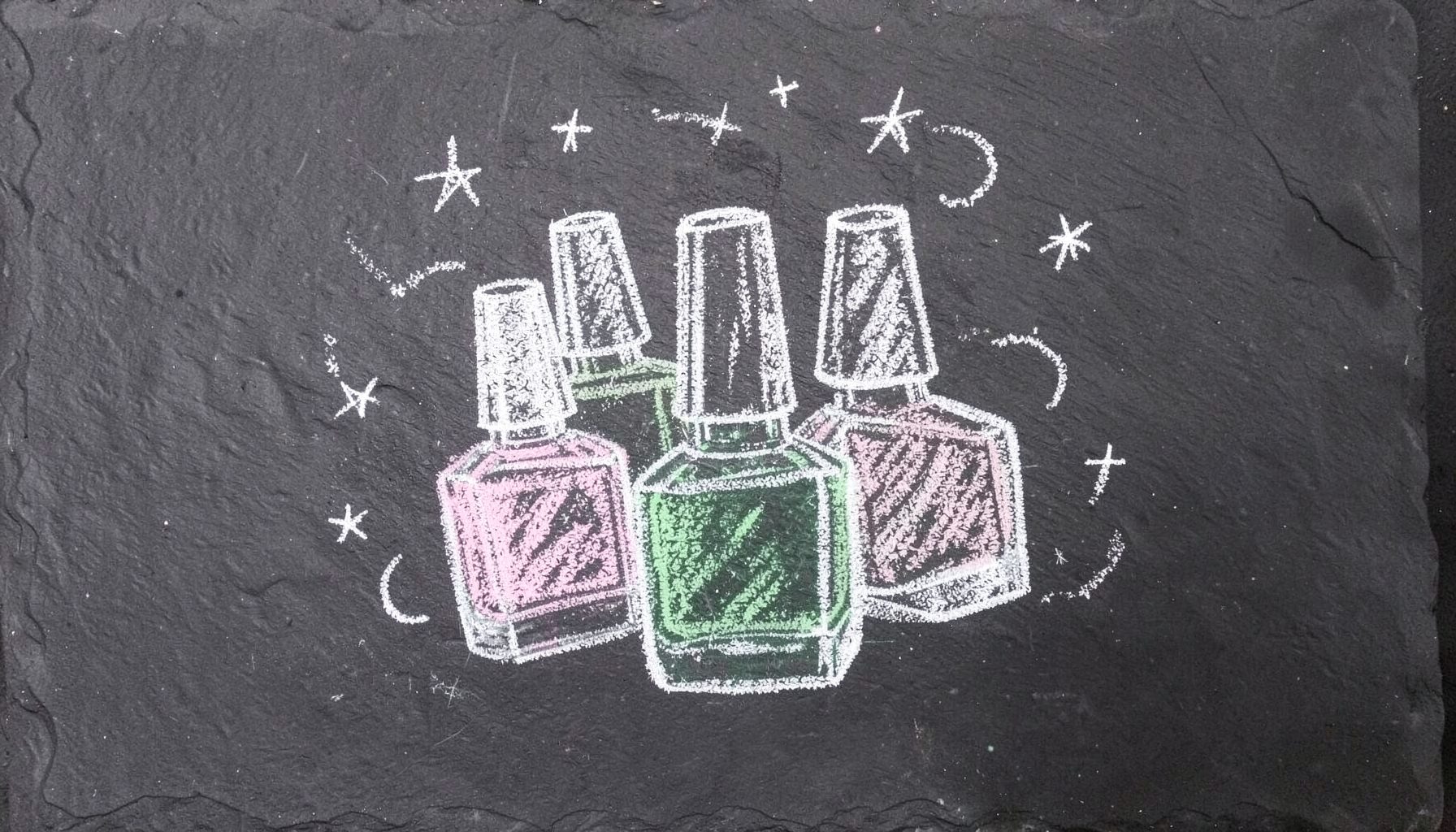 A chalk drawing on a dark slate board showing a cluster of nail polish bottles sketched in white and pastel chalk, with small stars and swirls around them, evoking a cosy handmade celebration.