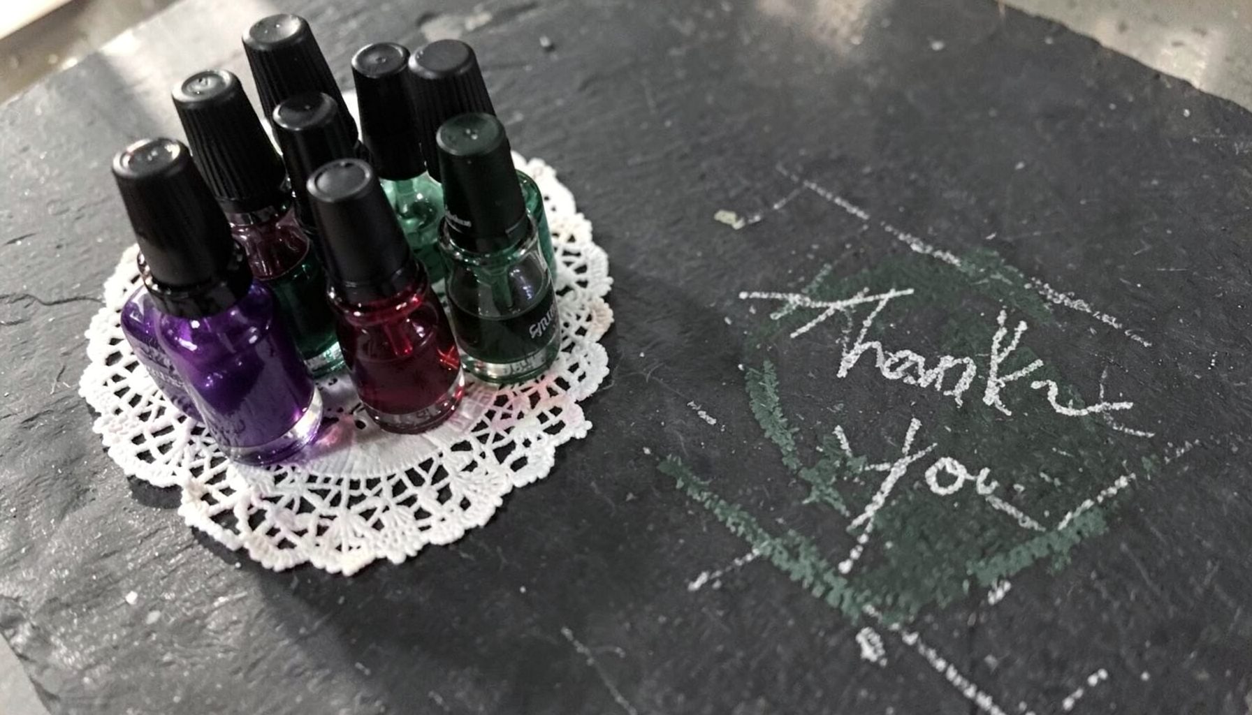 A cluster of small glass nail polish bottles in jewel tones — deep purple, emerald green, and ruby red — arranged on a white lace doily beside a handwritten 'Thank You' note, with soft natural light casting gentle shadows.