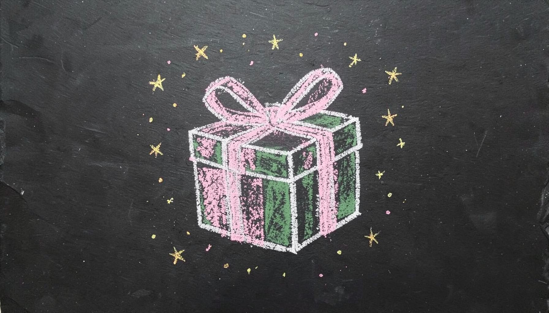 A chalk drawing on a dark slate board of a gift box tied with a ribbon, surrounded by tiny stars and confetti dots, drawn in soft pink and gold chalk.