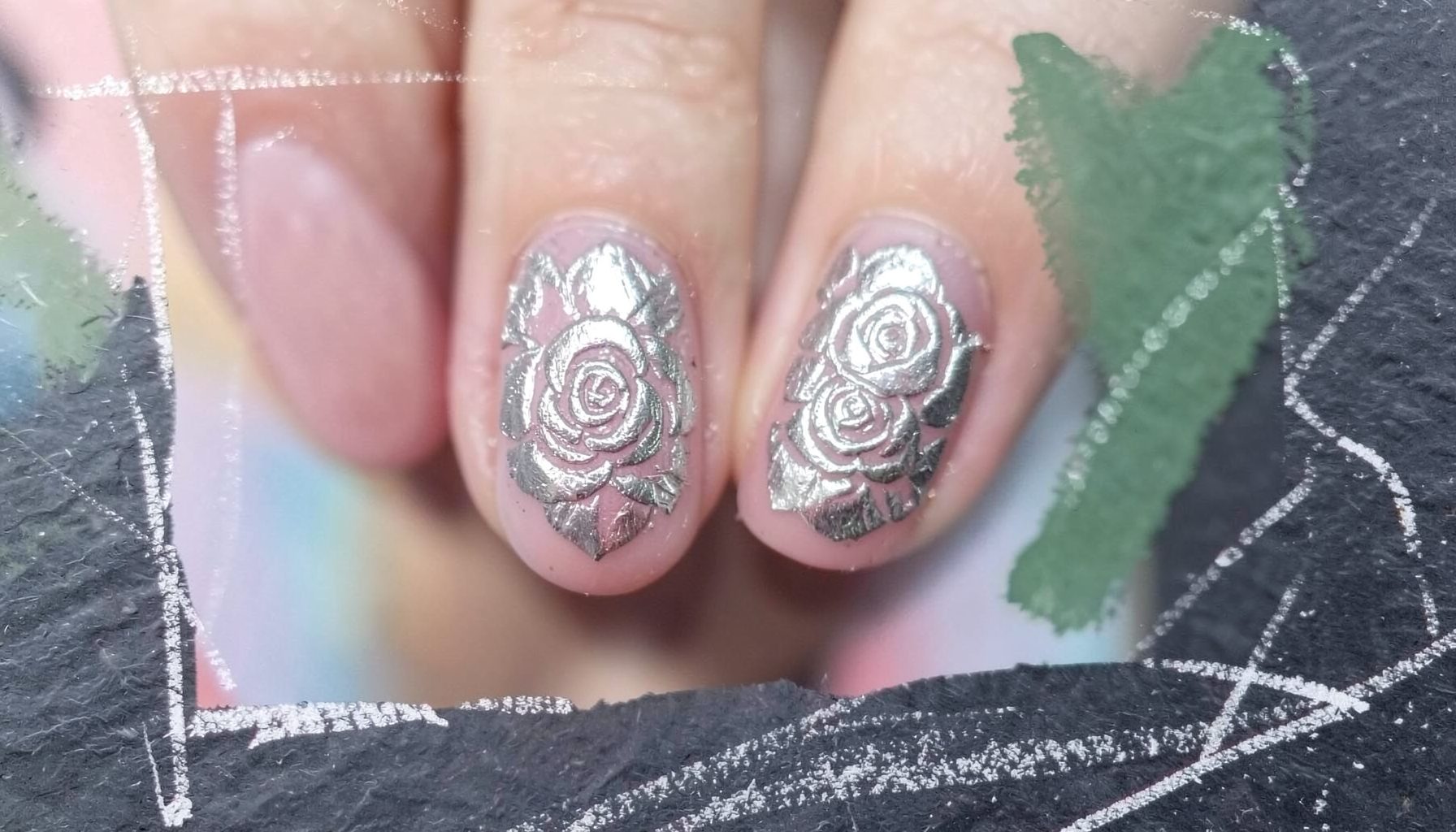 A close-up of fingernails stamped with an intricate rose pattern in metallic silver over a soft dusty-pink base, set against a blurred pastel background.